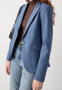 Blå skräddarsydd kavaj med satinfinish, slagkrage, enkel knappstängning, med en metallogo på fickan, bärs över en brun topp och jeans.