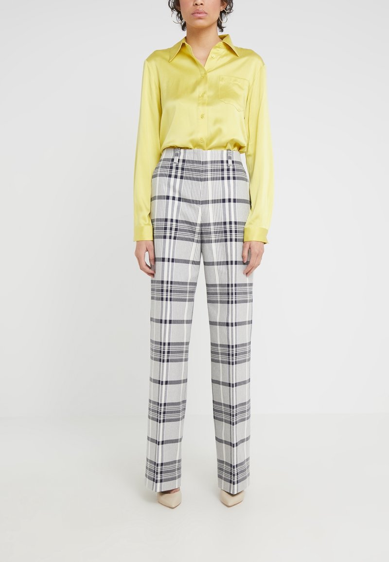 Personne portant une chemise jaune à manches longues rentrée dans un pantalon à carreaux blanc et noir taille haute et des chaussures beiges à bouts pointus, debout devant un fond uni.