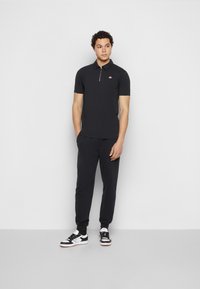 Čierne polo tričko s krátkymi rukávmi a predným zipsom, v kombinácii s čiernymi joggermi. Model má na sebe čierno-biele tenisky. Jednoduchý dizajn.