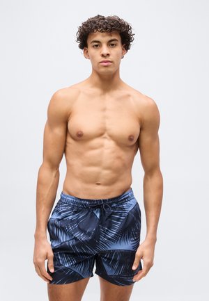 Mladý muž s kučeravými vlasmi v modro-čiernych vzorovaných plavkach, stojaci na jednoduchom svetlom pozadí.