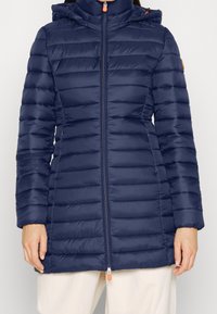 Tummansininen puhvitakki, jossa on vaakasuuntainen tikkauskuvio, korkea kaulus, vetoketjukiinnitys ja säädettävä huppu. Varustettu oranssilla vetoketjun vetimellä ja logolla.