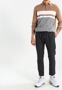 Polo a maniche lunghe marrone con strisce bianche e beige, abbinato a pantaloni tapered a righe nere e sneakers bianche. Texture liscia, design aderente.