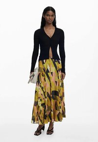 Schwarzer gerippter Cardigan mit tiefem V-Ausschnitt, kombiniert mit einem gelb-pink-schwarz gemusterten Maxirock, gehalten mit einer weißen Clutch und Sandalen.