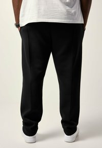 Man staat met zijn handen in zijn zakken, gekleed in zwarte broek, witte sneakers en een wit shirt tegen een effen lichte achtergrond.
