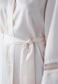Robe beige chiaro realizzato in tessuto morbido, con cintura annodata in vita e delicata rifinitura in pizzo sulle maniche. Texture morbida e fluida.