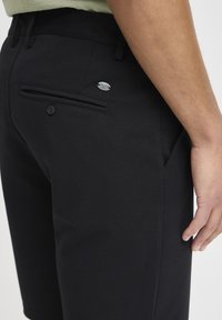 Black shorts made of woven fabric, featuring a back pocket with button and metallic logo accent. Smooth texture and tailored fit.