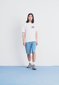 Hvit kortermet t-skjorte med et lite grafisk design på venstre bryst, kombinert med lyseblå denimshorts og grå sneakers.
