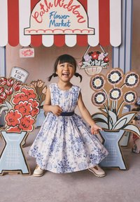 Jauna mergina mėlynose gėlėtoje suknelėje šypsosi ir stovi spalvingų kartoninių gėlių ekspozicijų apsuptyje Cath Kidston gėlių turguje.