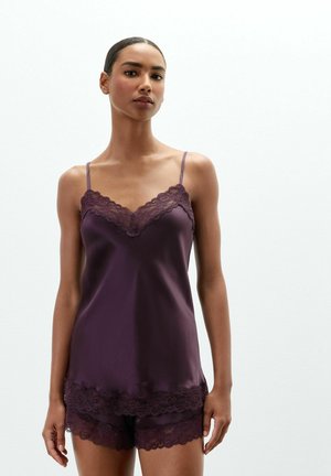 Kvinde iført mørkelilla satin camisole og matchende shorts med blonder står foran en ensfarvet hvid baggrund.