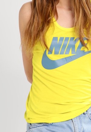 Person iført en gul Nike tanktop uden ærmer med blå logo og lyseblå denimjeans foran en ensfarvet baggrund.