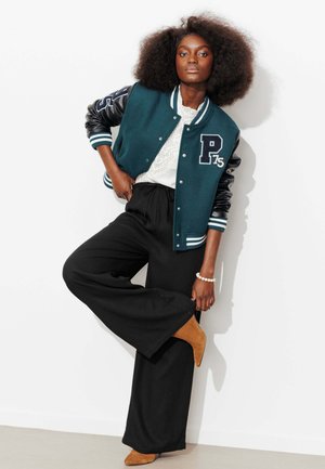 Femme avec une grande afro appuyée contre un mur blanc, portant une veste universitaire verte avec des manches en cuir noir, un pantalon large noir, un top blanc et des bottes marron.