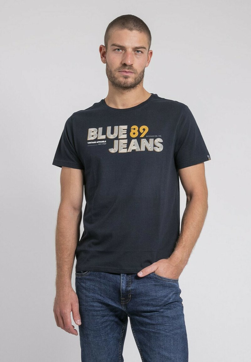 T-shirt en coton bleu marine à col rond, avec un graphique "BLUE 89 JEANS" en blanc et jaune. Associé à un jean bleu foncé.