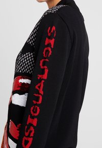 Svart tröja med röda paljetter i form av läppar och röd vertikal text "DESIGUAL" på ärmen, bärs av en person som står vänd åt sidan.