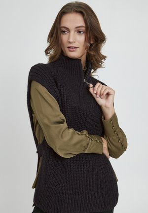 Dunkler gestrickter Weste mit hohem Kragen und Reißverschlussdetails, getragen über einem olivgrünen Langarmshirt mit Knopfakzenten. Strukturierter Stoff.
