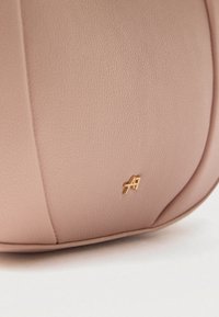 Κοντινή λήψη ροζ τσαντάκι από δέρμα με υφή, με μικρό χρυσό μεταλλικό λογότυπο στην κάτω μπροστινή πλευρά, σε λευκό φόντο.