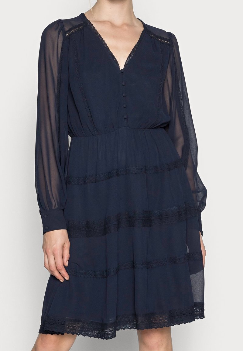 Robe bleu marine à manches longues avec manches en voile transparent, col en V, boutons sur le devant, détails en dentelle et jupe à volants.