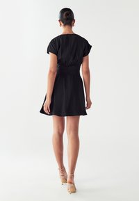 Čierne šaty s krátkymi rukávmi a priliehavým pásom, s rozšírením na leme. Hladká tkanina, viditeľný zips na chrbte. Spárované s telovými lodičkami.