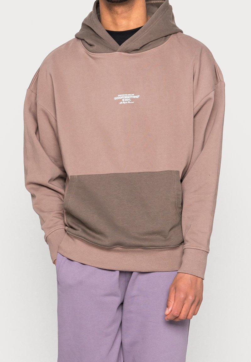 Homme portant un sweat à capuche marron bicolore avec poche avant et pantalon lavande, debout avec une main dans la poche et l'autre détendue.