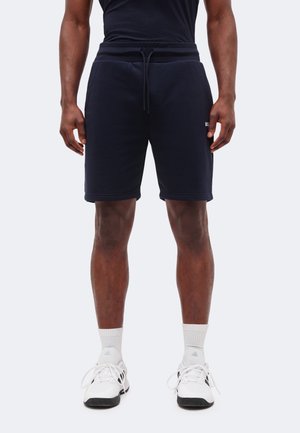 Άνδρας μοντέλο φοράει σορτς σε σκούρο μπλε με κορδόνι, λευκές κάλτσες μέχρι τον αστράγαλο και λευκά αθλητικά παπούτσια Adidas, στέκεται μπροστά σε απλό φωτεινό φόντο.