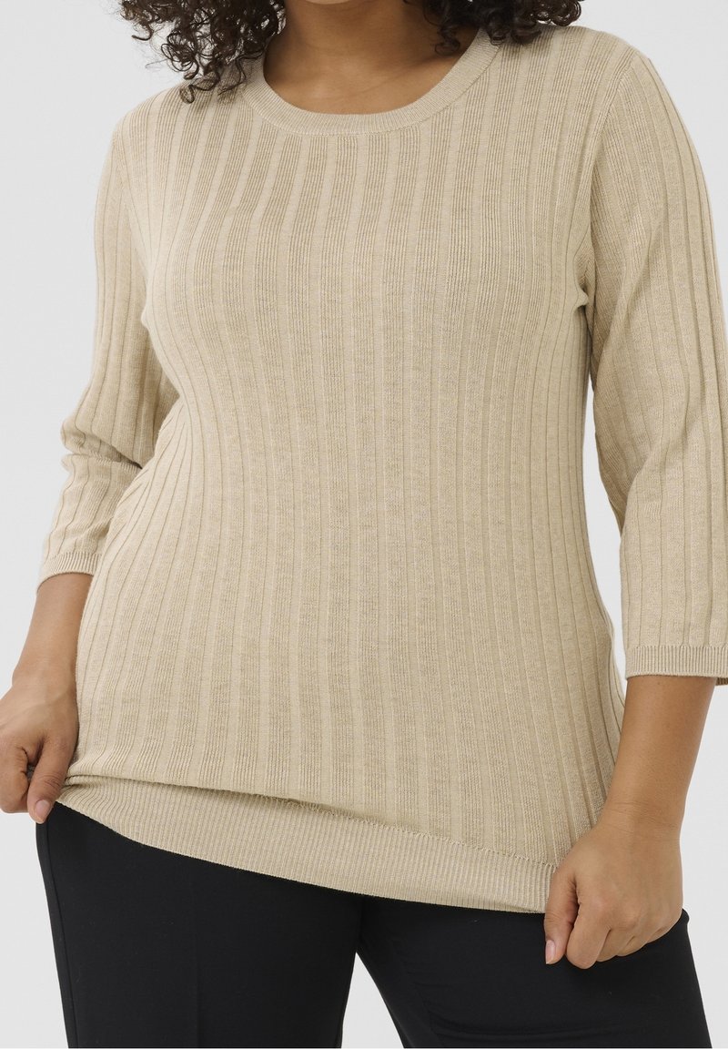 Femme portant un pull côtelé beige à manches trois-quarts et un pantalon noir, tenant l'ourlet du pull d'une main.