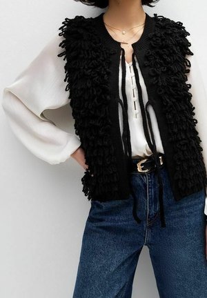 Femme portant un chemisier blanc à manches longues avec un gilet sans manches noir texturé, un jean bleu et un collier fin superposé.