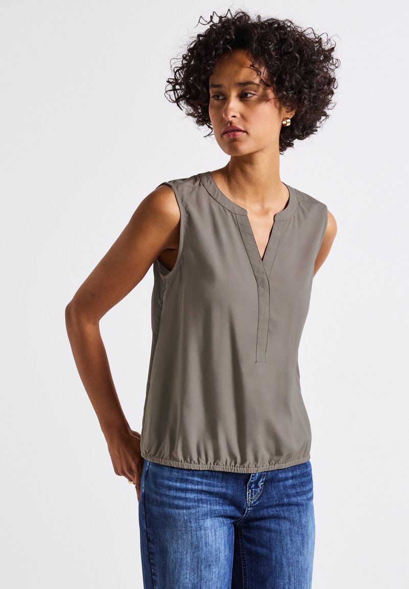 Ärmellose Bluse in Olivgrün, mit V-Ausschnitt und gerafftem elastischen Saum. Glatter, leichter Stoff mit minimaler Detailgestaltung. Kombiniert mit blauen Jeans.