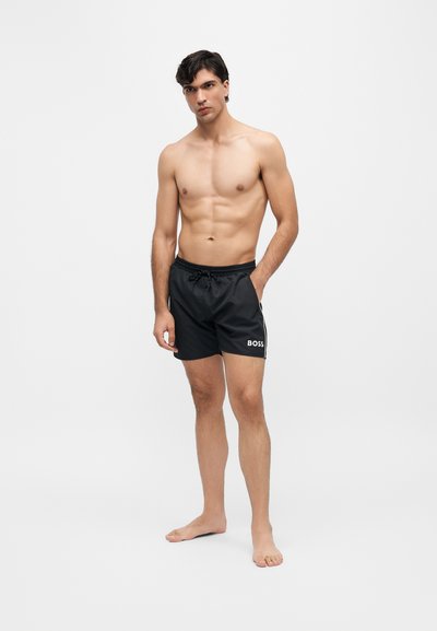 Jeune homme debout pieds nus, portant un short de bain noir avec le logo "BOSS" sur la jambe gauche, regardant devant lui avec une main dans la poche.