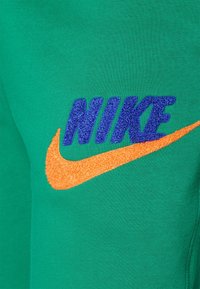 Mikina Nike; zelená látka s hladkou texturou. Obsahuje modré vyšívané nápis "NIKE" a oranžové texturované logo ve tvaru šipky.