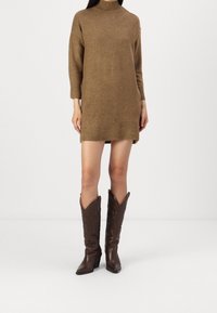 Abito in maglia marrone con colletto alto e maniche a tre quarti, abbinato a stivali da cowboy marroni scuri con dettagli in rilievo.