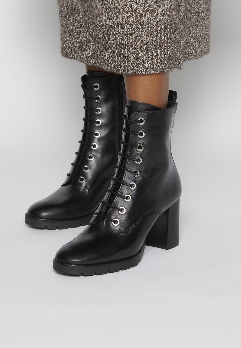 Botas de tobillo de cuero negro con cordones y tacones de bloque, combinadas con una falda de punto marrón, sobre un fondo gris claro.