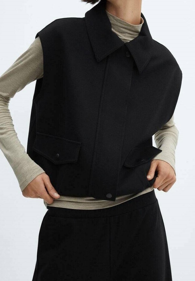 Personne portant une veste noire sans manches par-dessus une chemise beige à manches longues, les mains reposant près des poches sur un fond uni.