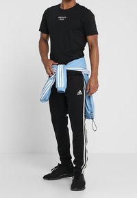 Czarna koszulka Manchester United, czarne dresowe spodnie Adidas z białymi paskami, niebieska kurtka związana w talii, czarne adidasy.