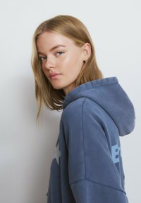 Sudadera con capucha azul con un diseño destacado de una estrella blanca, tela texturizada y capucha con cordón, usada por un modelo posando de lado.