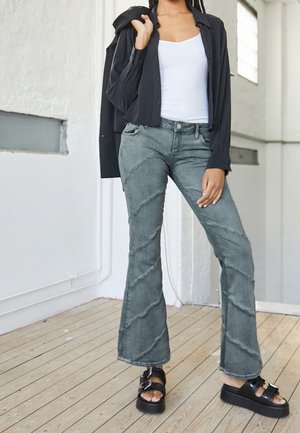 Person står indendørs iført en hvid tanktop, sort åben skjorte båret over skulderen, grå udvidede jeans og sorte platformsandaler.