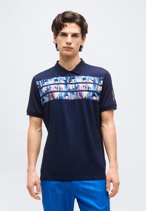 Νέος άνδρας με κοντά καστανά μαλλιά, φορώντας μπλε σκούρο πόλο με πολύχρωμες αφηρημένες ρίγες στο στήθος και μπλε σορτς με σχέδιο.