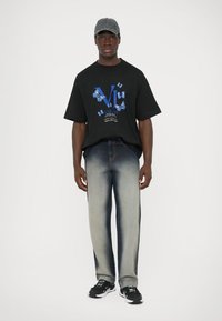 T-shirt oversized preta com estampa de borboleta azul e logótipo. Jeans de gradiente azul claro e ténis pretos. Boné cinza completa o visual.