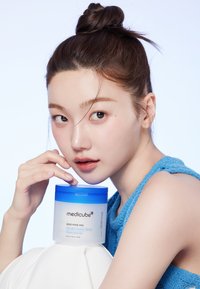 Medicube Zero Pore Pad-behållare, rund och transparent, med ett blått lock. Modell med blått texturerat topp, ljus hudton och naturlig makeup.