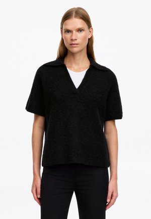 Femme portant un pull noir duveteux à manches courtes avec col et encolure en V sur une chemise blanche, debout devant un fond blanc uni.