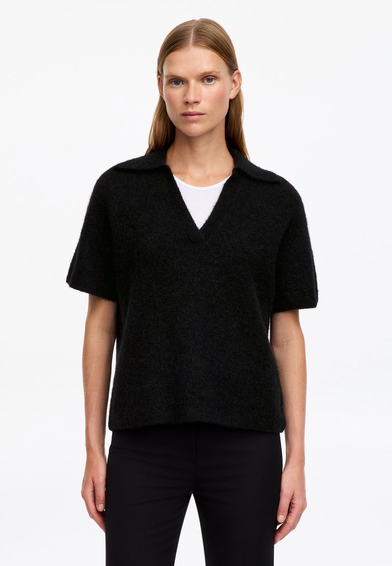 Femme portant un pull noir duveteux à manches courtes avec col et encolure en V sur une chemise blanche, debout devant un fond blanc uni.