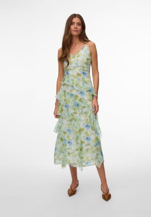 Kvinde iført en ærmeløs, lagdelt blomstret kjole med blå og grønne mønstre, parret med brune spidse hæle mod en hvid baggrund.