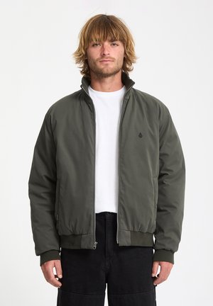 Dark green bomber jacket with a zip front, high collar, and ribbed cuffs. Worn over a white t-shirt, features a small logo on the chest.
