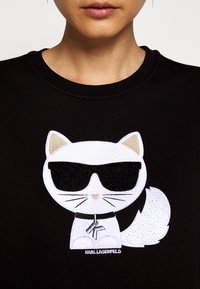 Femme portant un t-shirt noir avec un patch de chat dessin animé blanc, portant des lunettes de soleil noires et un collier avec un pendentif « K ».