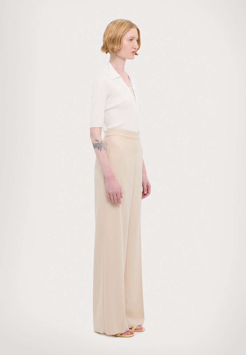 Top bianco a maniche corte con effetto costina e dettagli con lacci, abbinato a pantaloni beige a gamba larga, caratterizzati da una trama liscia e un design a vita alta.