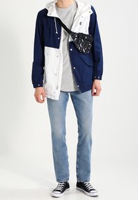 Rompevientos bloqueado en color azul marino y blanco con capucha, combinado con una camiseta gris, jeans azul claro y zapatillas negras. Bolso cruzado con pequeño estampado.