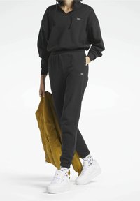 Osoba v černé mikině a teplácích, v bílých teniskách a vzorovaných ponožkách, držící v ruce hořčicově žlutou bundu.