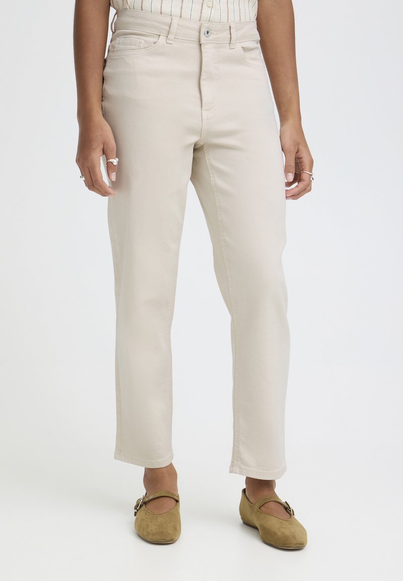 Beežid sirged kõrge vöökohaga püksid, mida kannab seisev isik koos pruunide seemisnahast Mary Jane stiilis kingadega.