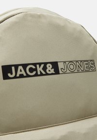 Mochila de tela beige con una superficie texturizada, que presenta un prominente logo negro que dice "JACK & JONES" en la parte superior. Diseño simple y redondeado.
