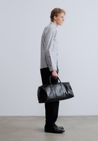 Μαύρη υφασμάτινη τσάντα duffel με δερμάτινες λαβές, με υφαντό σχέδιο. Άνδρας με γκρι πουκάμισο και μαύρο παντελόνι στέκεται κρατώντας την τσάντα.