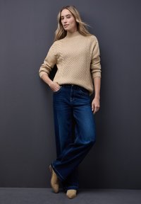 Beżowy sweter w warkocze z wysokim dekoltem, zestawiony z szerokimi granatowymi dżinsami. Modelka nosi jasnobrązowe botki za kostkę.