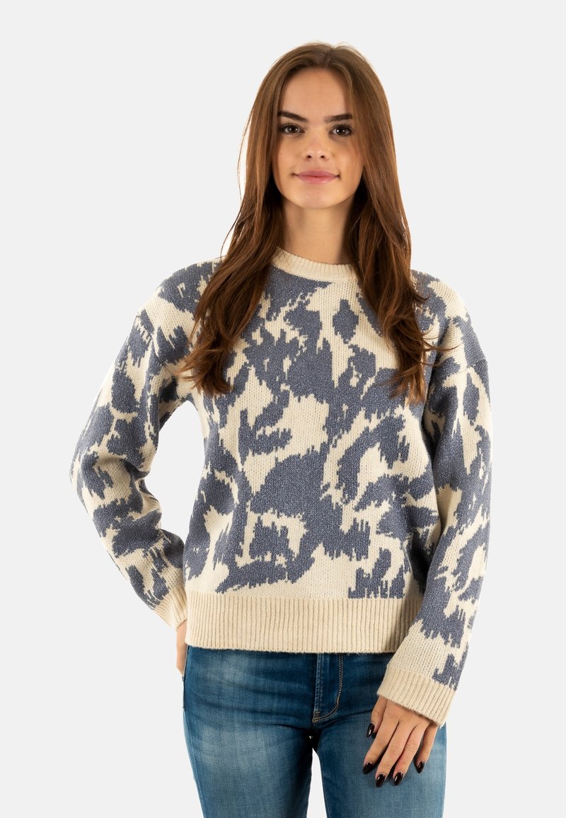 Pull en ivoire avec un motif abstrait bleu ; tissu tricoté doux, encolure ronde, poignets et ourlet côtelés. Associé à un jean bleu.